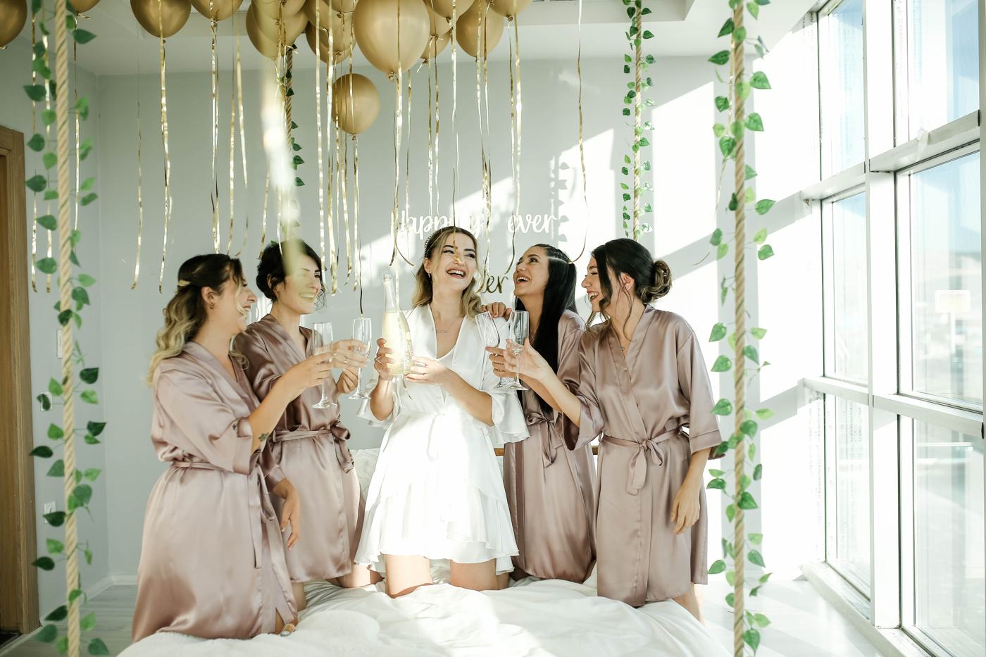 A bride and four bridesmaids in robes raise champagne glasses