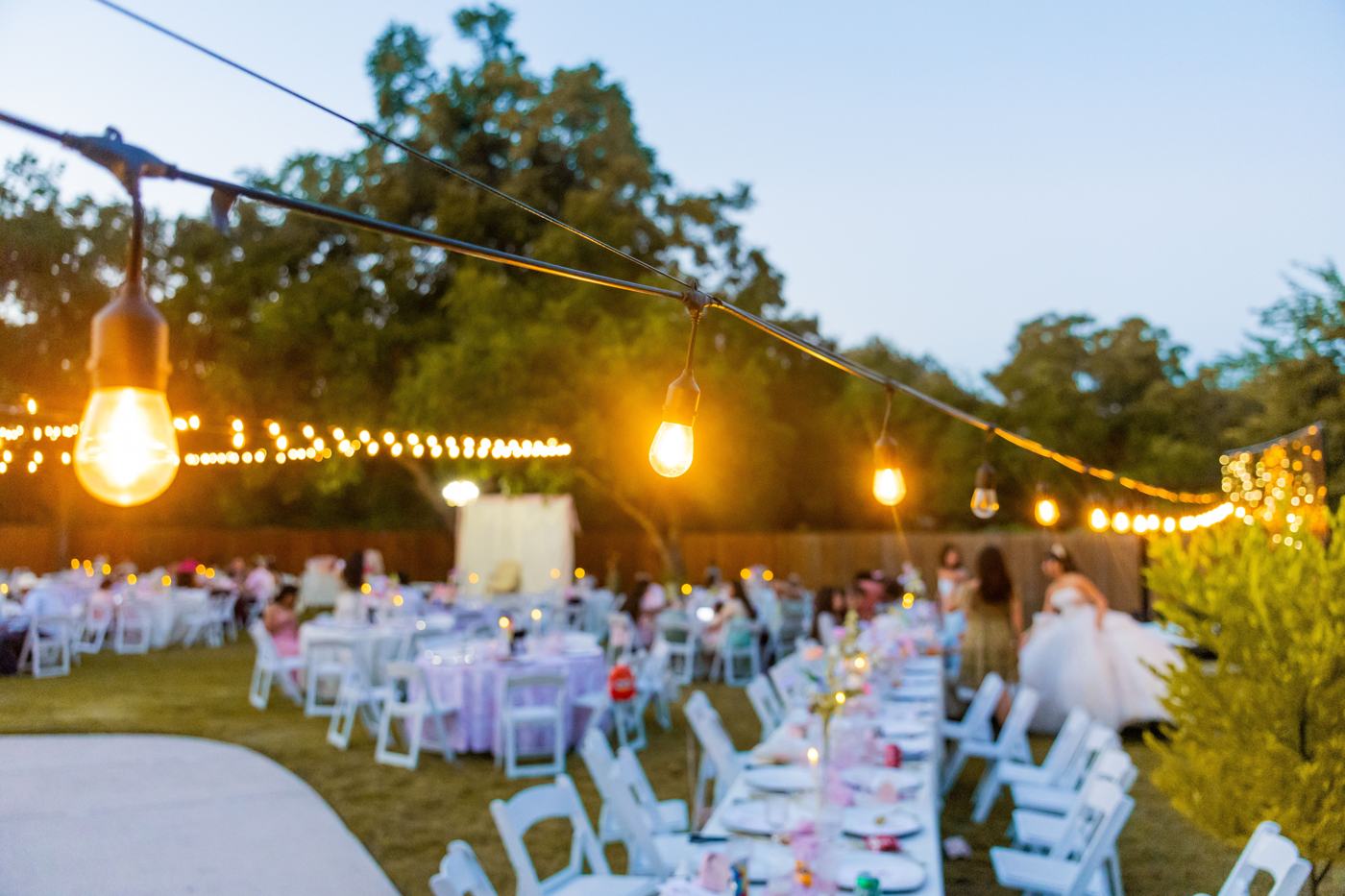 Long tables set under string lights at an outdoor wedding reception at dusk
